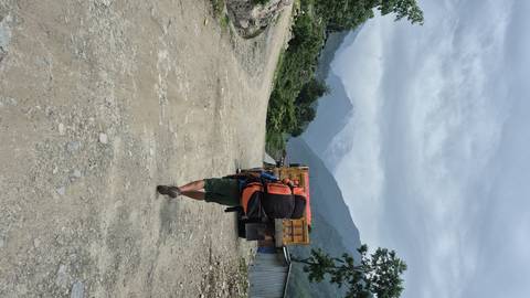       Person with large backpack walking on a dirt road, mountains in view.
  