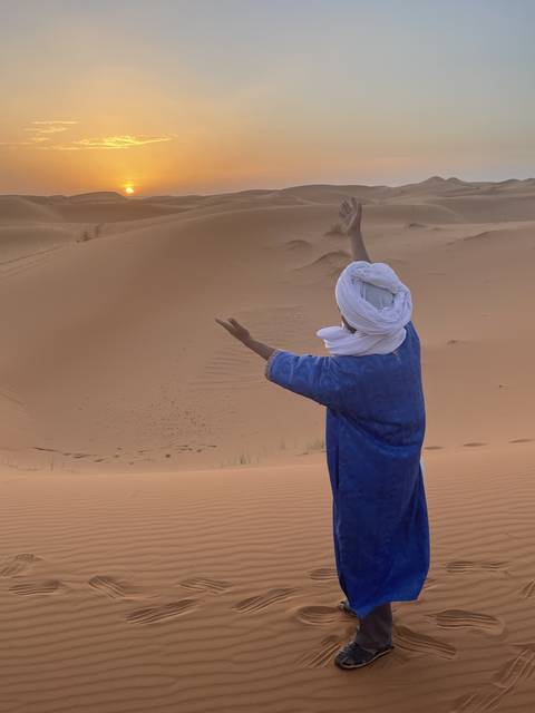       Person in traditional clothing looking at a sunset over sand dunes.
  