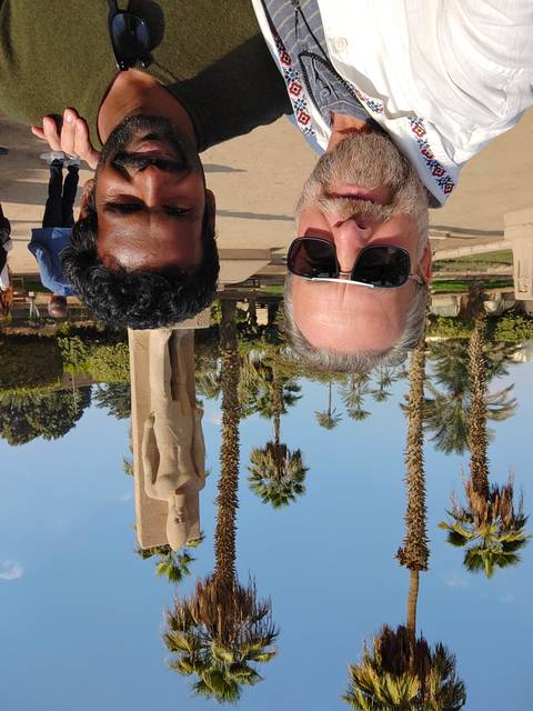 Two men taking a selfie near palm trees and a statue.