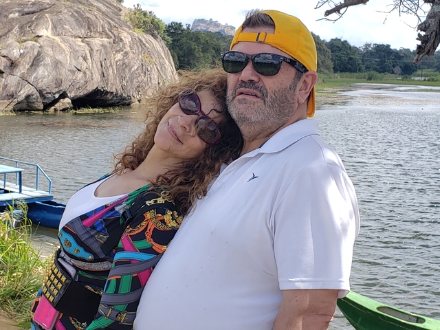       Couple posing by a lake with a boat in the background.
  