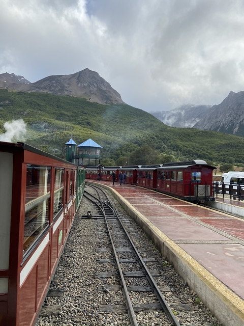       Train station with red train and forested mountains.
  