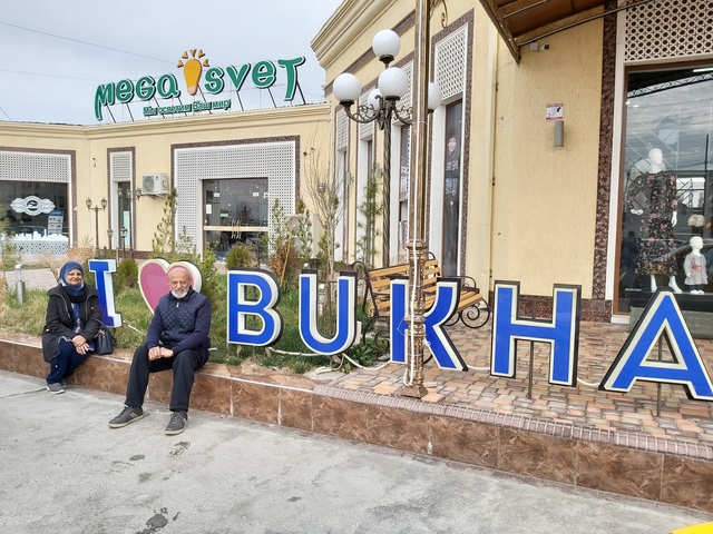Two people seated in front of a sign that says 'I Love Bukhara'.