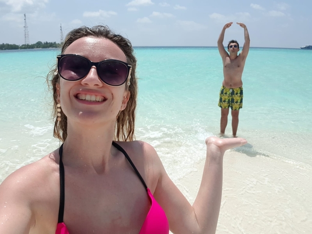 A woman taking a selfie with a man playfully posing in the background at the beach.