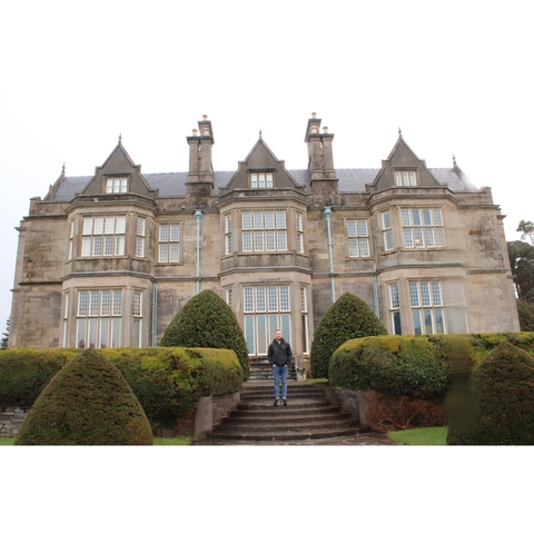 Person standing in front of a large stone mansion with greenery.