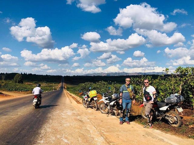 Motorcyclists standing beside bikes on a long road with clear skies.