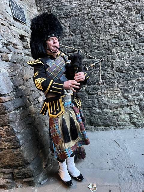 Bagpiper in a traditional outfit playing in front of a stone wall.