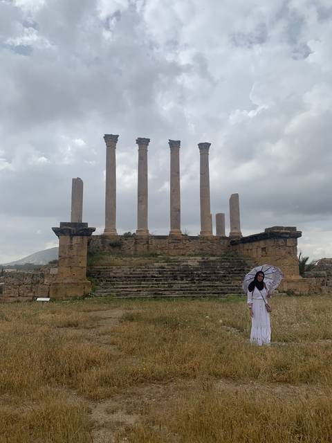       Person posing in front of ancient ruins with columns.
  