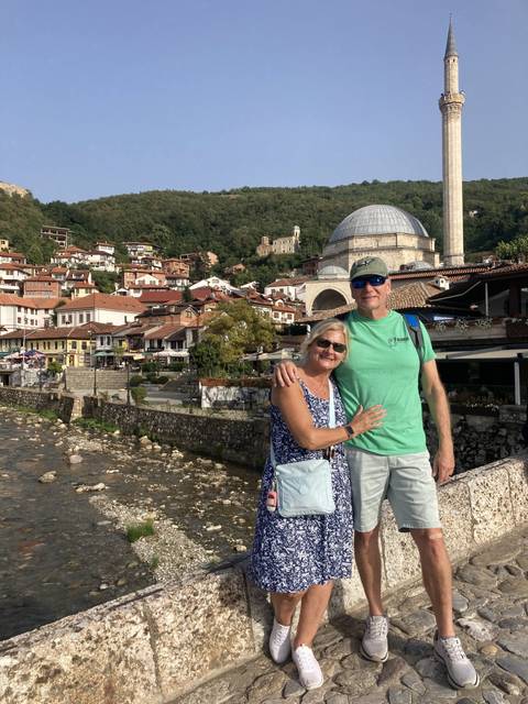 Two people posing on a bridge with a view of buildings and a river.