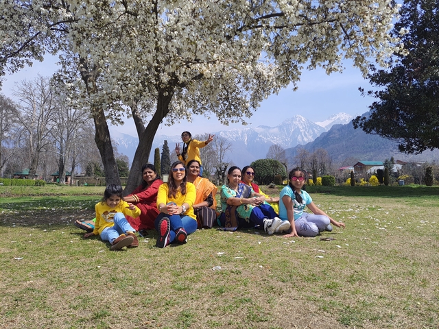 Group of people seated under a flowering tree with mountains behind.