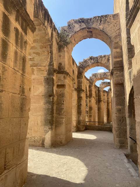       A stone corridor with large arches and sunlight casting shadows.
  