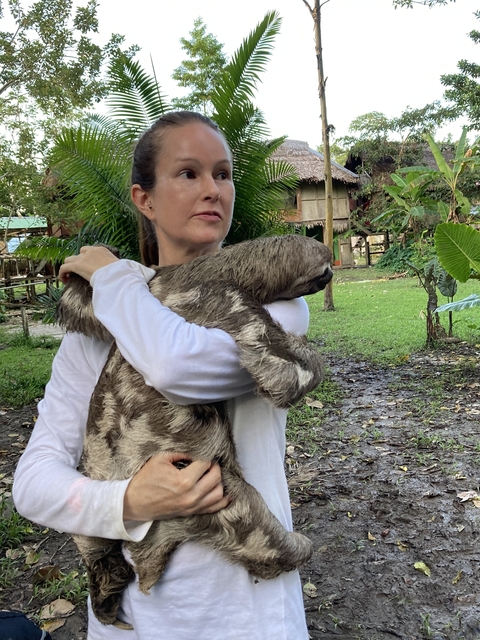 Woman holding a large sloth in an outdoor area with greenery.