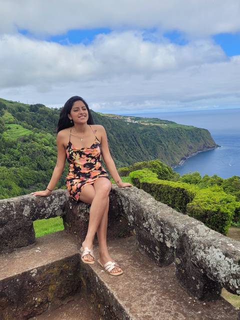 Woman sitting on a stone wall with scenic views of green hills and the sea.