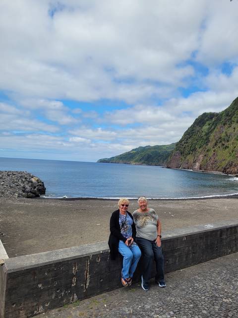       Couple sitting on a bench by the ocean with hills in the background.
  