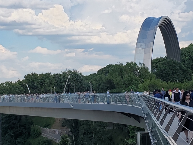 Crowded pedestrian bridge with a large arch in the background.