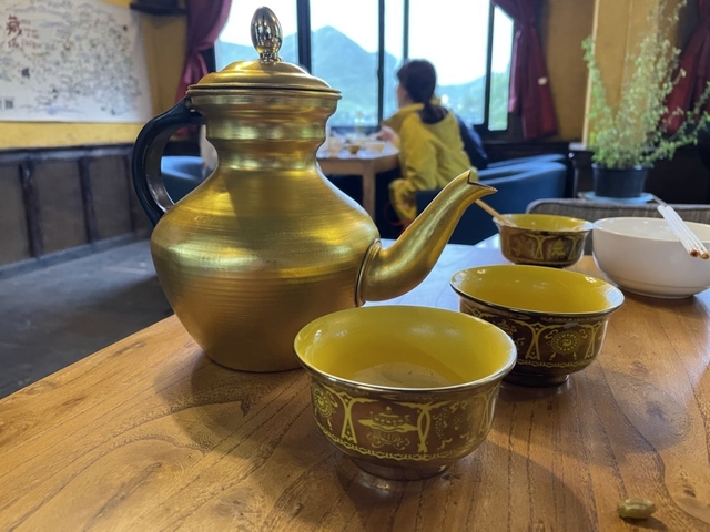 Golden teapot with two cups on a table and people in the background.
