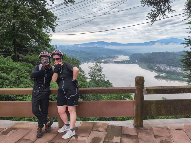 Two cyclists posing with a scenic view of a river and hills.