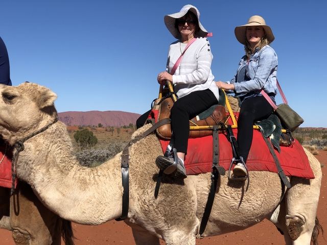       Two people riding a camel with desert landscape and red soil.
  