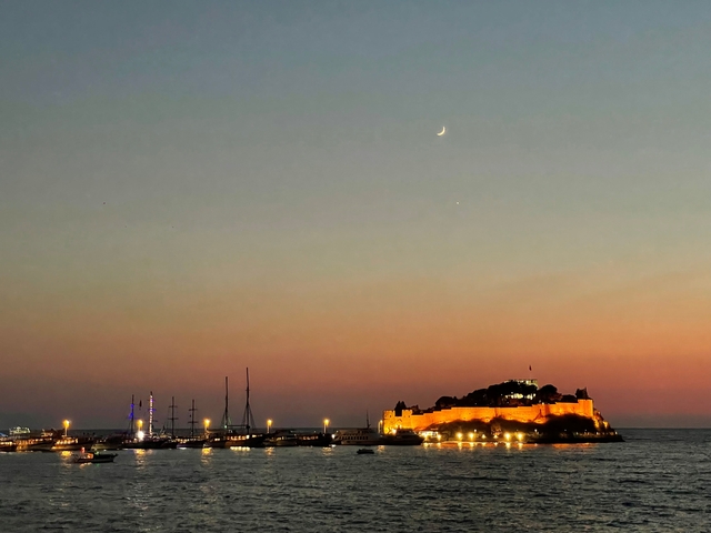 Seaside fort lit up at sunset with crescent moon.