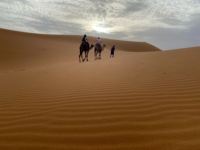 Camel riders in the desert under a cloudy sky.