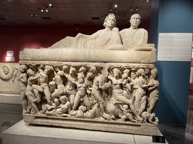       Sarcophagus with detailed sculptures surrounded by a museum interior.
  