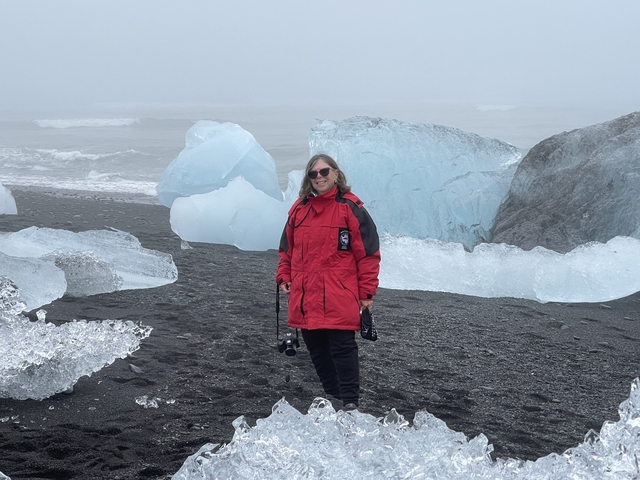 Person in red coat standing on a black sand beach with icebergs.