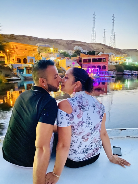       A couple facing each other by a river with colorful lights in the background.
  