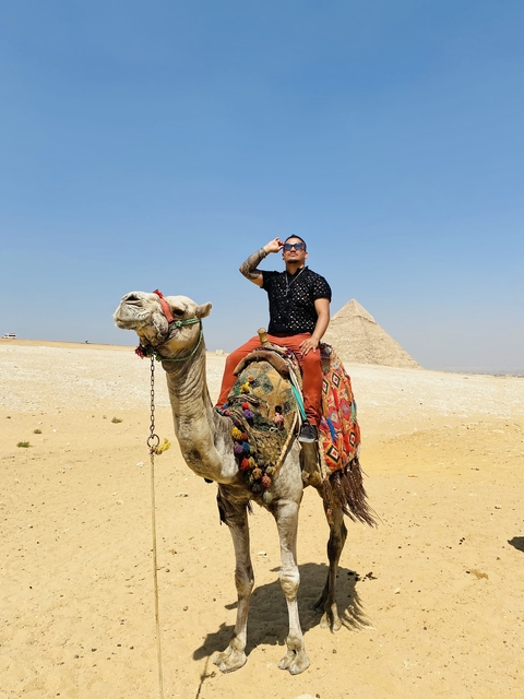       Man riding a camel with pyramids in the background.
  