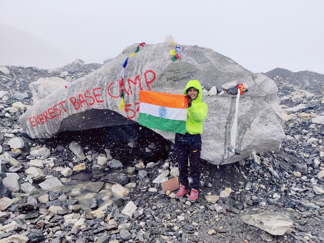       Person at Everest Base Camp holding a flag next to a marked rock.
  