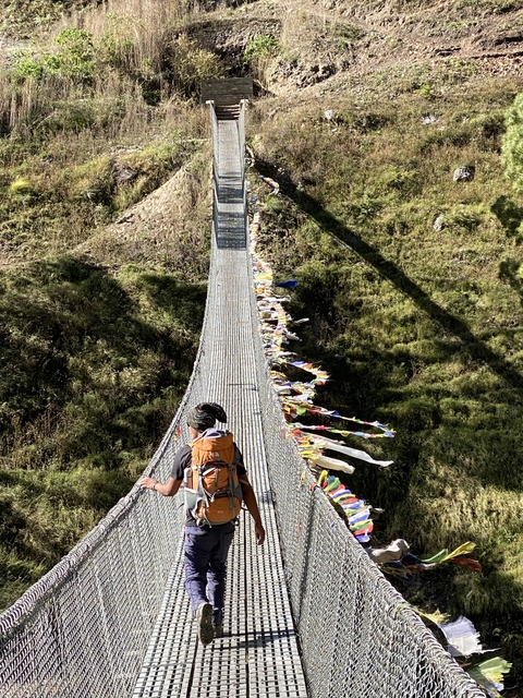       Person crossing a hanging bridge adorned with prayer flags in a hilly terrain.
  