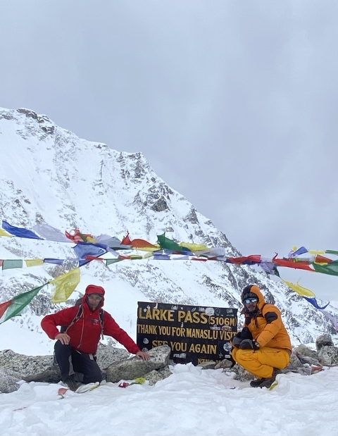       Two people at a snowy mountain pass with colorful flags.
  