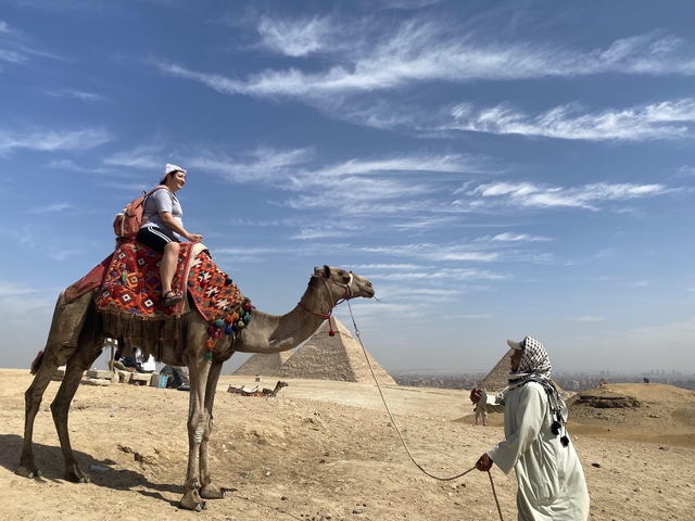Person riding a camel with pyramids in the background.