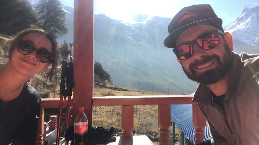 Two people smiling with trekking poles; snowy mountains in the background.