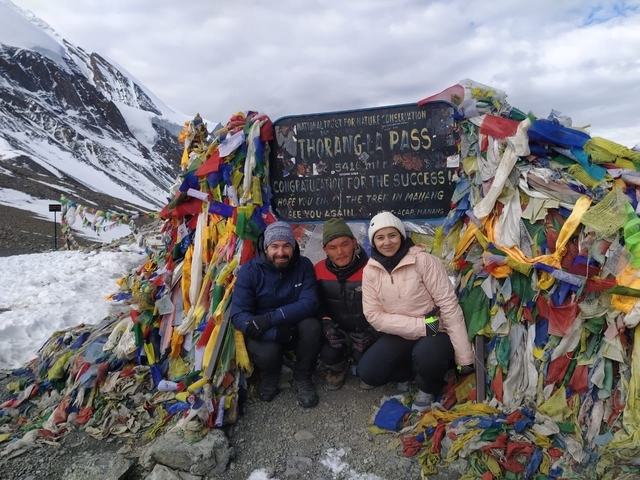 Three people posing at Thorong La Pass with colorful prayer flags and a snowy mountain backdrop.