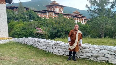       A person in traditional Bhutanese attire standing with a backdrop of architecture.
  