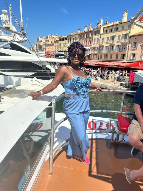 Person posing on a boat with colorful buildings and water in the background.