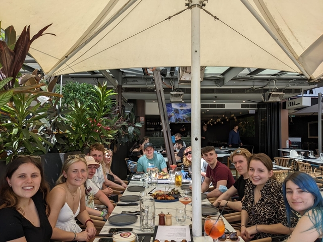       A group of people dining outdoors under an umbrella.
  