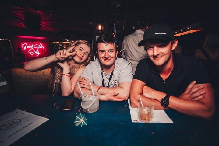 Three people at a bar with drinks, neon sign in background.