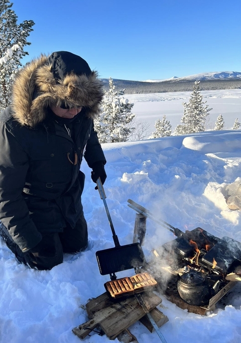 Person in winter clothing cooking over an open fire in the snow.