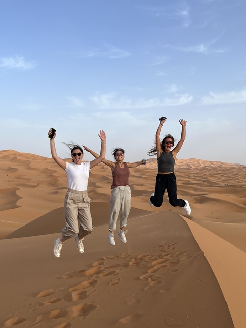 Three women jumping in a desert with dunes.