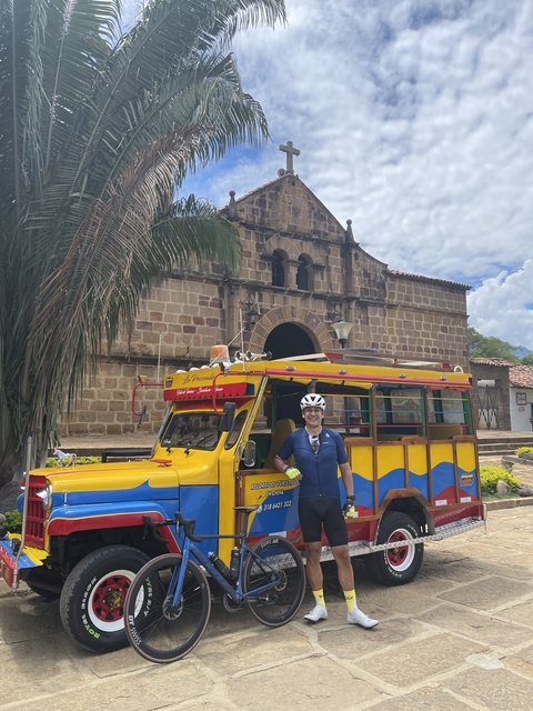 Cyclist standing in front of a colorful traditional vehicle with a church behind.