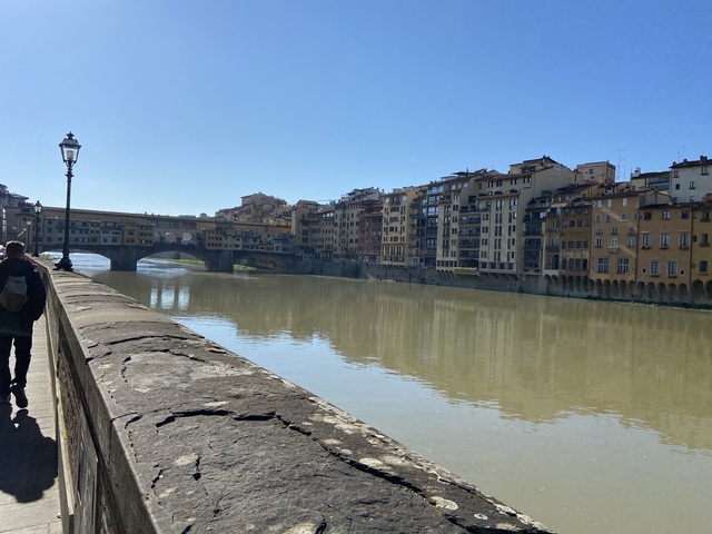 A person walking along a riverbank with historic buildings.
