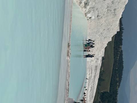       Tourists walking in shallow turquoise water near white travertine terraces.
  