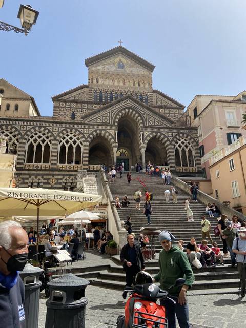      People walking up steps of a historic cathedral with intricate facade in a lively square.
  