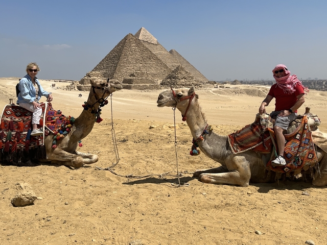       Two people on camels with pyramids in the background.
  