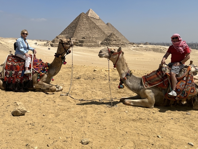       Two people on camels with pyramids in the background.
  