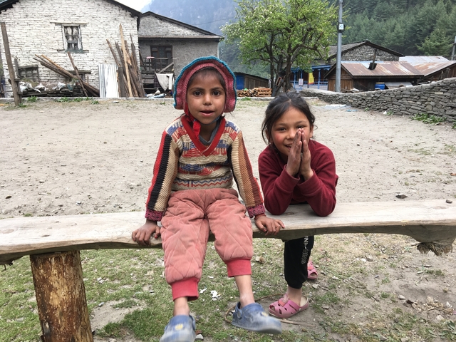       Two children sitting outside on a bench in a village.
  