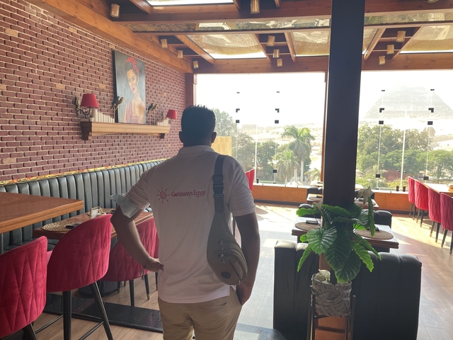 Interior of a restaurant with a person looking outside; pyramid visible through the window.