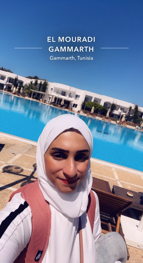       Selfie of a person in front of a swimming pool with a hotel in the background.
  