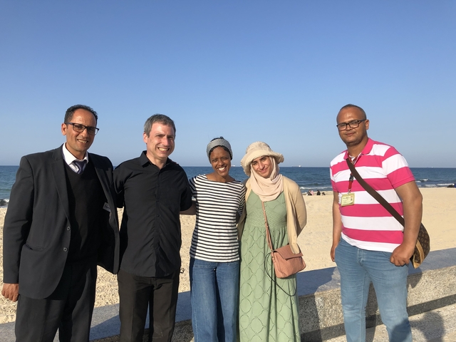       Group of five people posing on a beach with the ocean in the background.
  