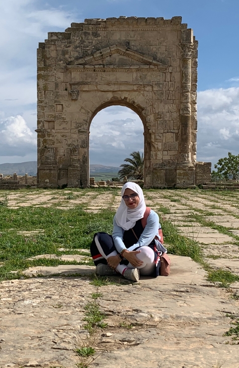       Person seated in front of an ancient arch with scenic surroundings.
  